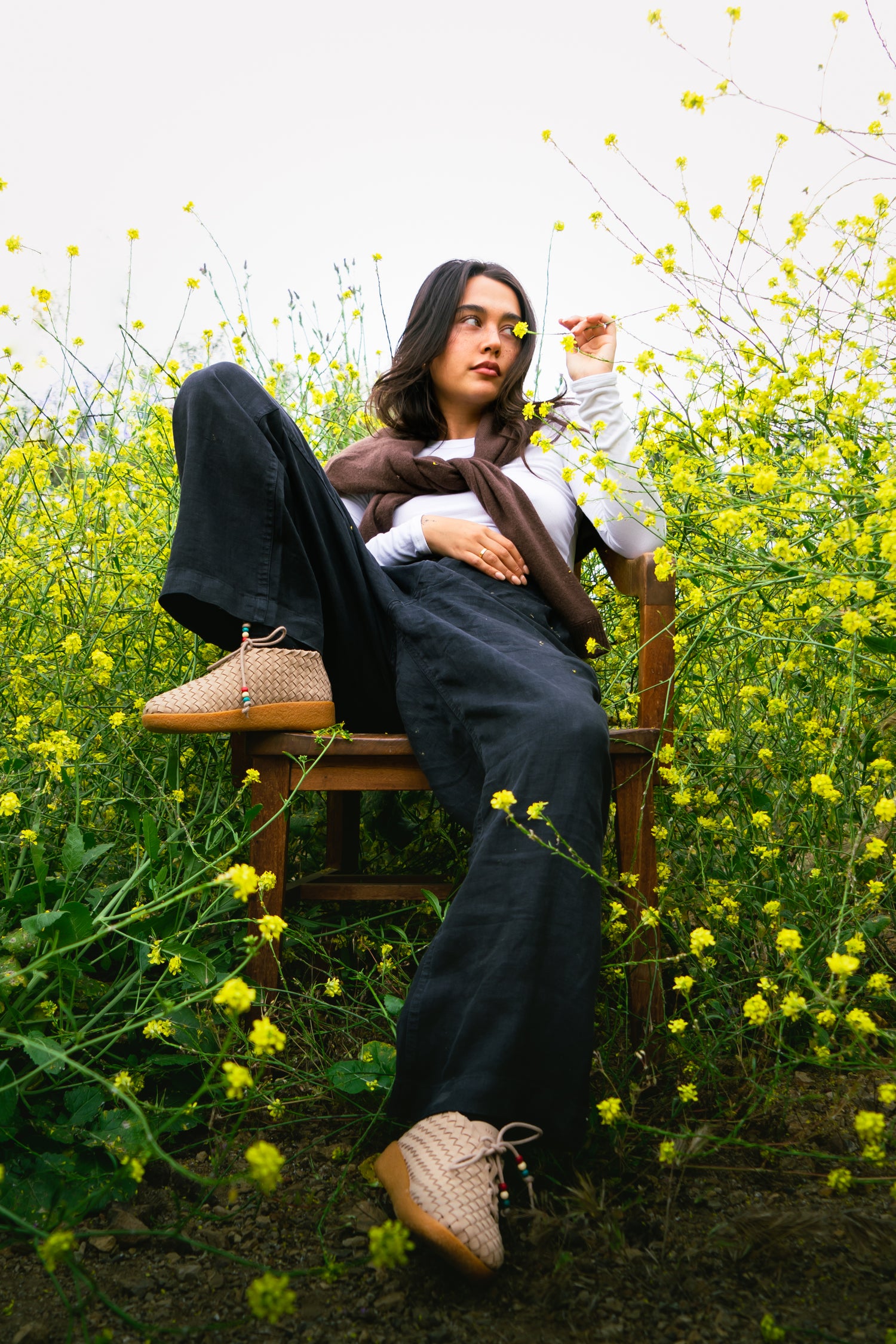 lifesyle photo shoot of a girl wearing the all new Malibu Sandals Matador chukka low wearing black pants, white long sleeve shirt and a brown sweater wrapped around her shoulders while surrounded by yellow flowers all around. 