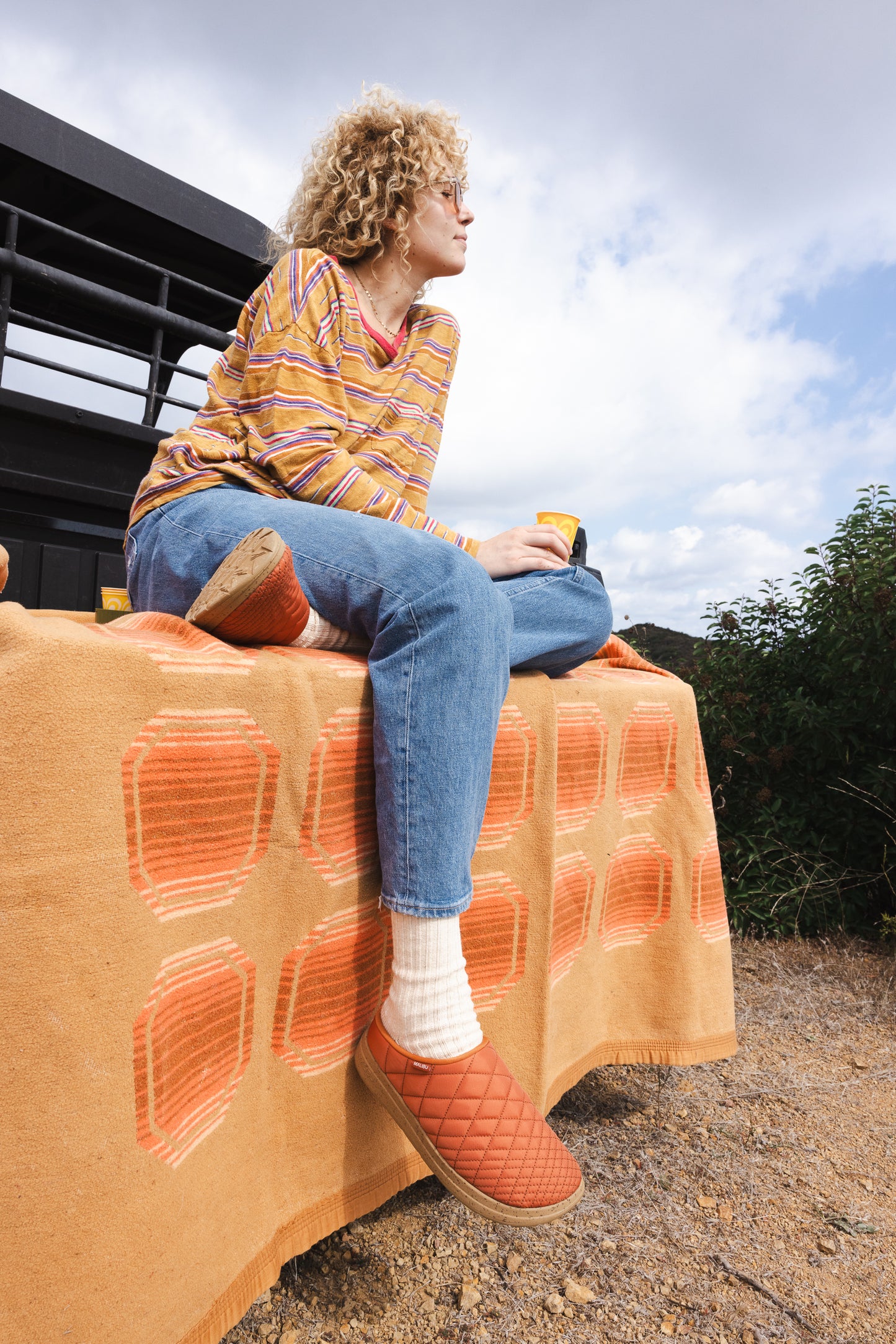 Lifestyle photo of a women sitting down looking in the distance while wearing the Malibu sandals westward ballistic clog in rust