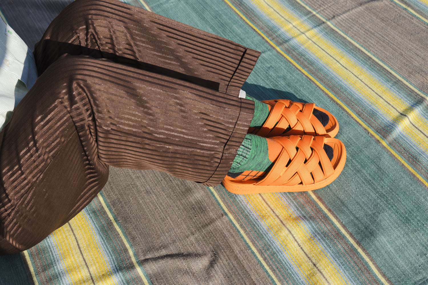 close up photo or person wearing Malibu Sandals Orange Zuma LX recycled poly slide with green socks brown stripped pants on a green and yellow stripped blanket