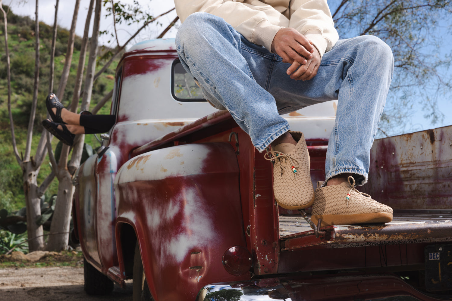 Lifestyle photo of Malibu Sandals Matador in beige being worn by a person sitting on the bed of a truck next to someone wearing the vegan leather Canyon. 