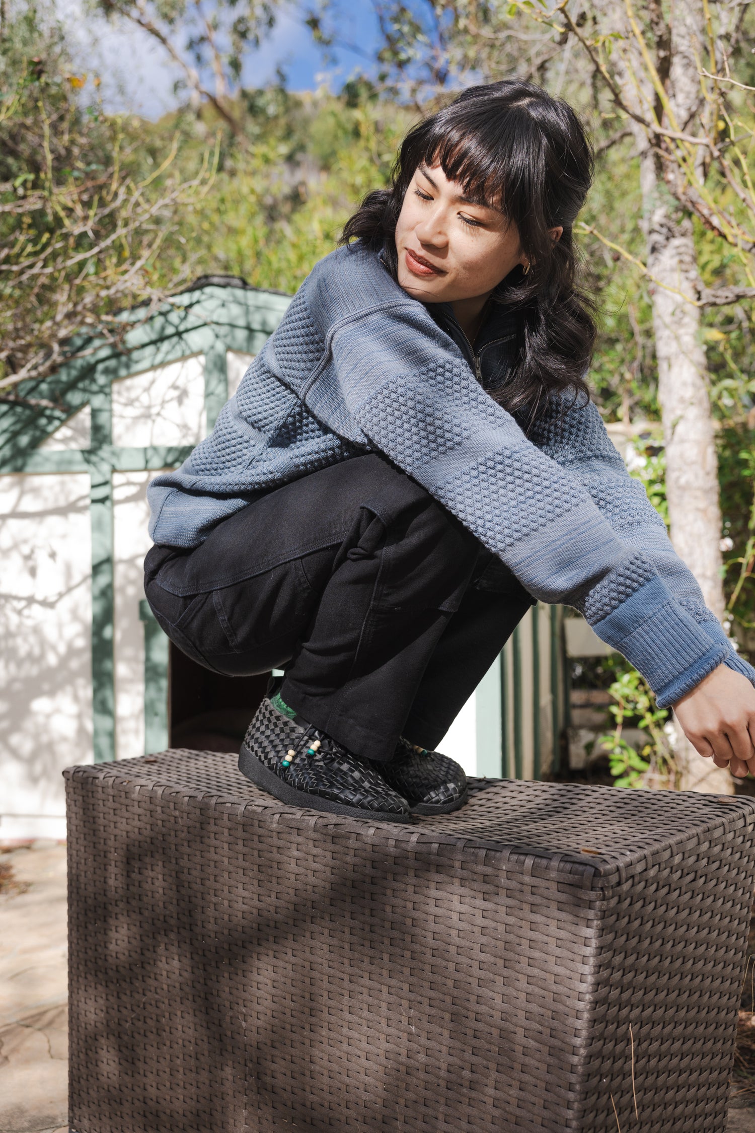 lifestyle photo of women with long black hair wearing Malibu Sandals all new Matador Chukka low in all black vegan leather with black pants and a faded blue sweater while crouching on a woven prop