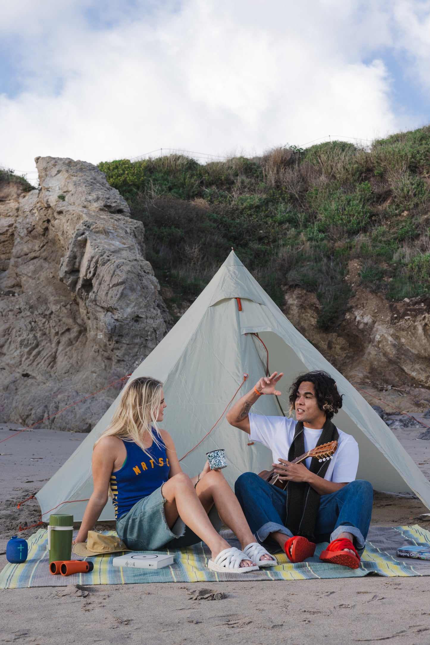 lifestyle photo of of a guy and girl in front of a tent sitting on a blanket on the beach while wearing Malibu Sandals all new Zuma LX recycled poly in white and red 