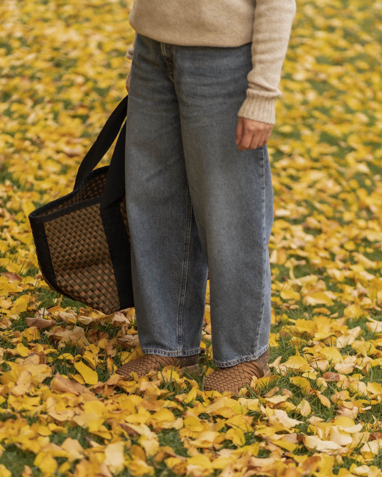 Image of a person standing on a bed of leaves holding Malibu sandals Weekender hand woven tote bag in olive black while also wearing the colony closed toe sandals in Coyote. 